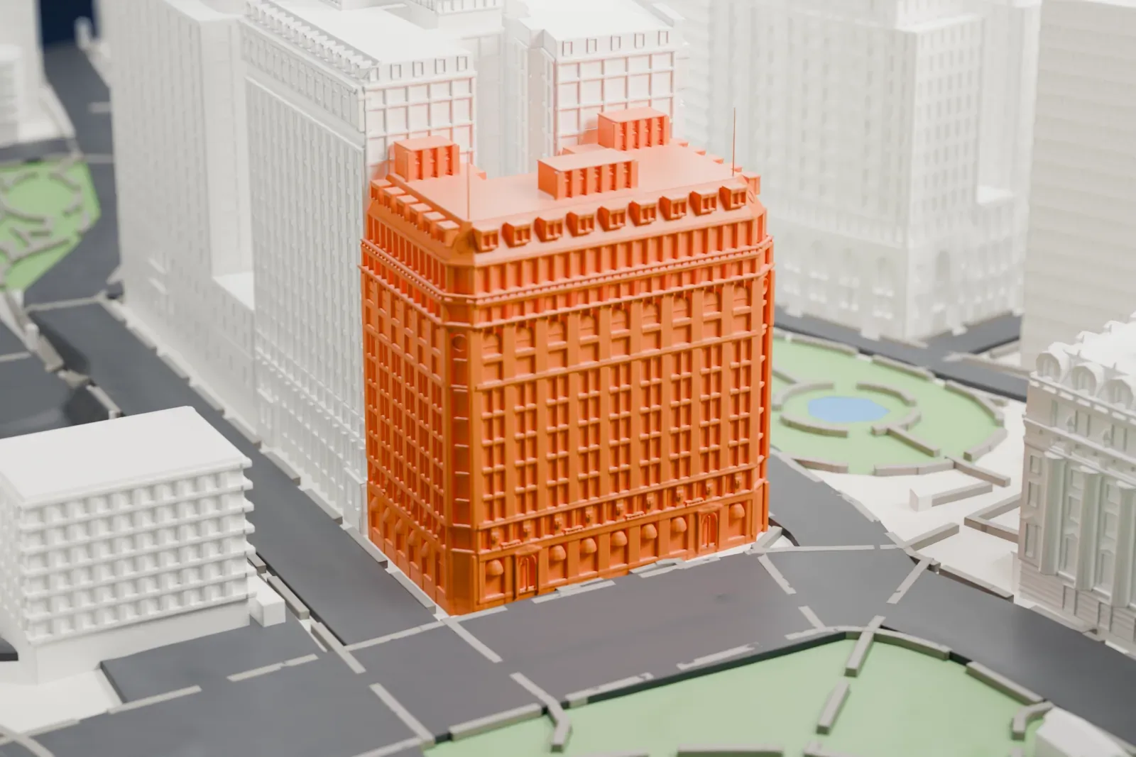 A white 3D print of 1 Broadway, a tall building with a dormered mansard roof and a few rooftop boxes, on a street corner beside a green park.