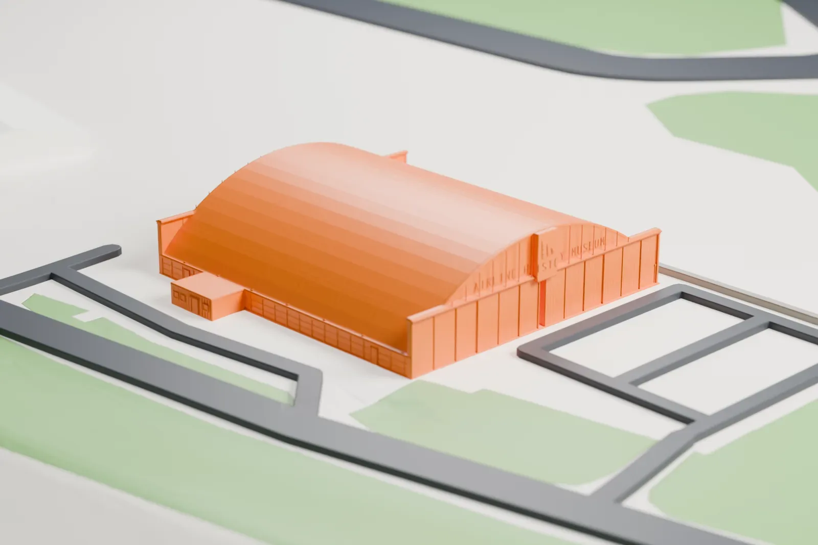 A white 3D print of Hangar 9, a long hangar with a curved barrel-vault roof, sitting on an open airport ramp with roads and grass around it.