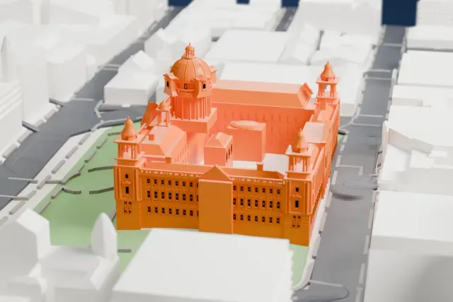 A white 3D print of Belfast City Hall, with its central dome and four corner towers, in green grounds with paths, ringed by streets and buildings.