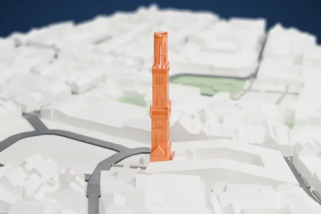 A white 3D print of the Belfry of Bruges, a tall bell tower with a pinnacled top, rising among dense low buildings and curving streets with a little water and green nearby.