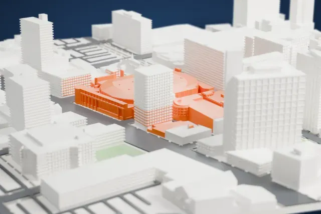 A white 3D print of Canada Life Centre, a low arena with a broad roof and a rounded corner, hemmed in by tall downtown towers and streets.
