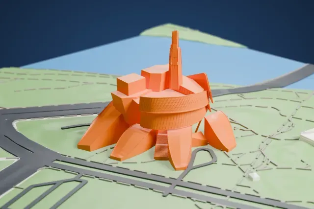 A white 3D print of the Canadian Museum for Human Rights, a cluster of angular wings around a tall thin spire, set on a green site with curving roads and water nearby.