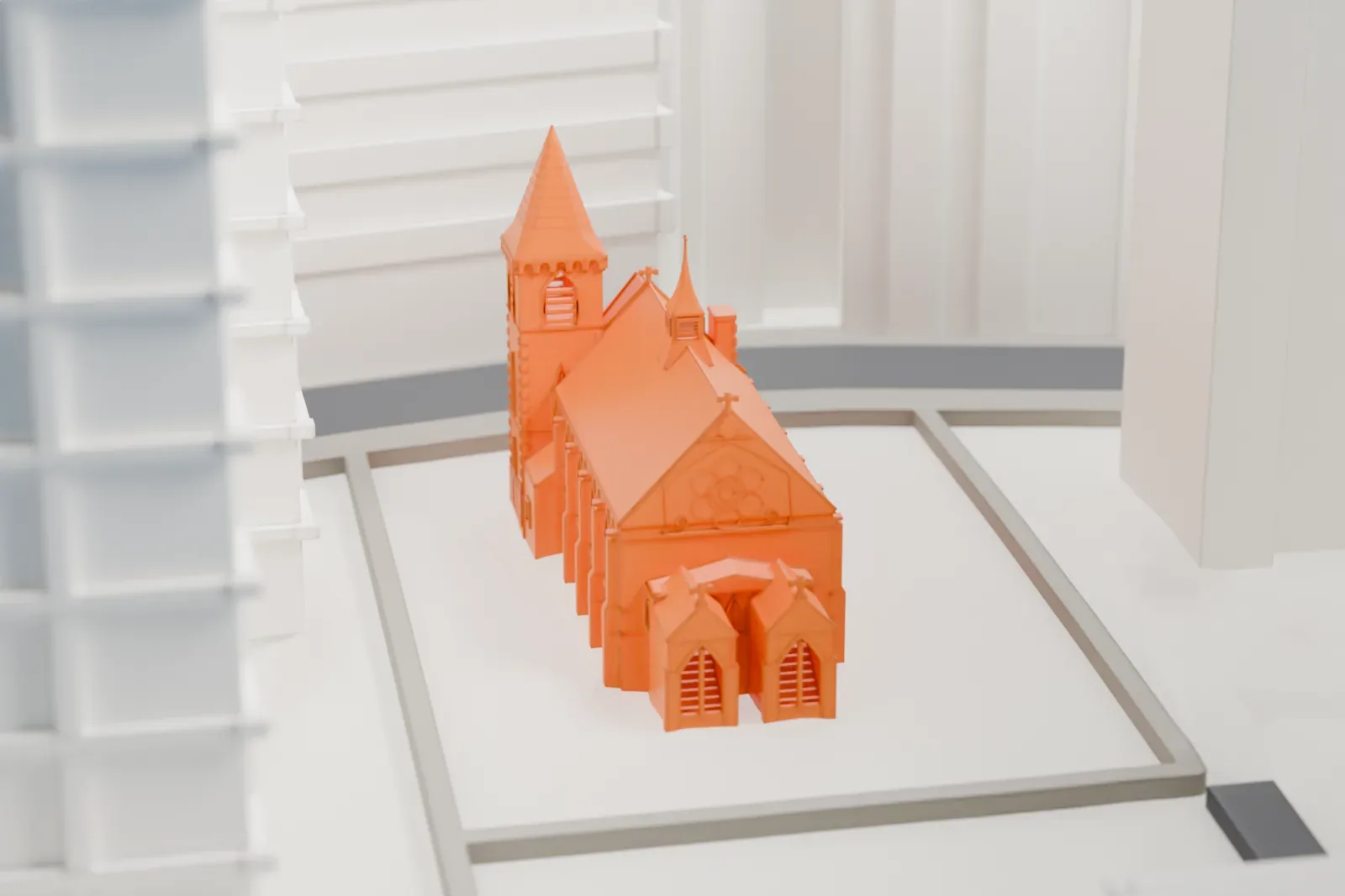 A white 3D print of the Chapel of the Good Shepherd, a steep roof with a small belfry and cross, set in a courtyard between taller buildings.