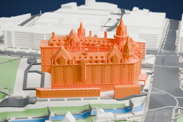 A white 3D print of the Château Laurier, with its turrets, dormers and steep roofs, beside a park and a strip of water with city buildings around it.