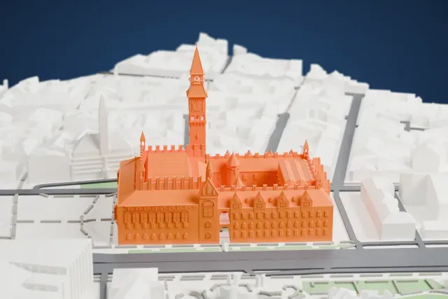 A white 3D print of Copenhagen City Hall, a large building with a tall slim clock tower, gabled roofs and small turrets, with water and city blocks around it.