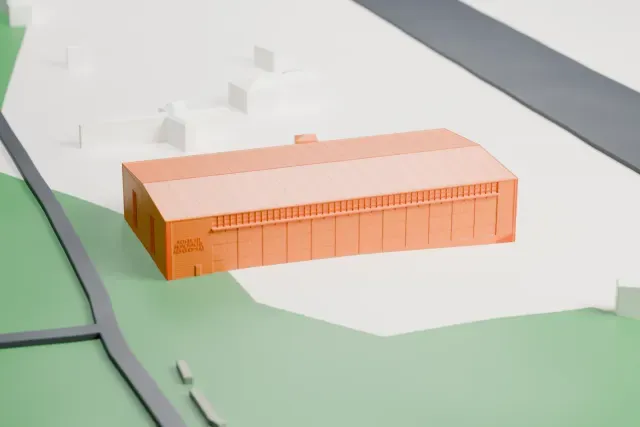 A white 3D print of the Hellenic Air Force Museum hangar, a large, low building with a ridged roof and small windows on its end walls, standing on open ground with a road and a few small buildings nearby.