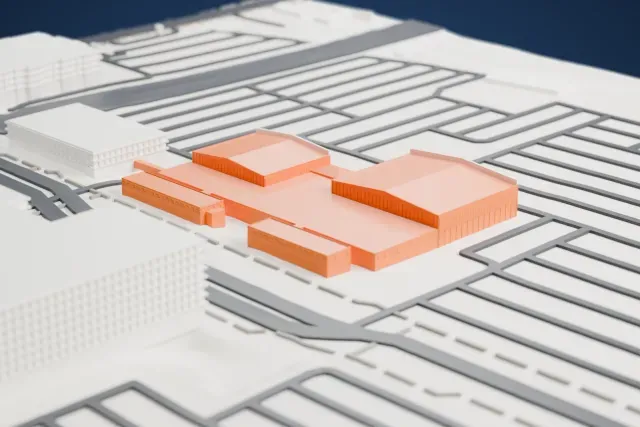 A white 3D print of the Delta Flight Museum hangars, two long gabled halls joined by a low building, surrounded by roads and a few other buildings.