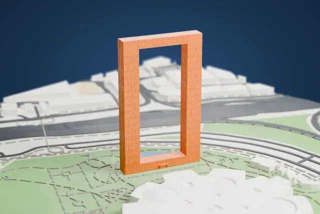 A white 3D print of the Dubai Frame, a tall rectangular frame with patterned relief faces, standing in a green park with paths and a small pond beside wide roads and a large domed building.