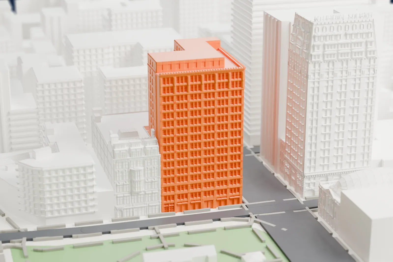 A white 3D print of the Everett Building, a tall rectangular block with a notched roof, standing beside a green park and surrounded by other tall buildings.