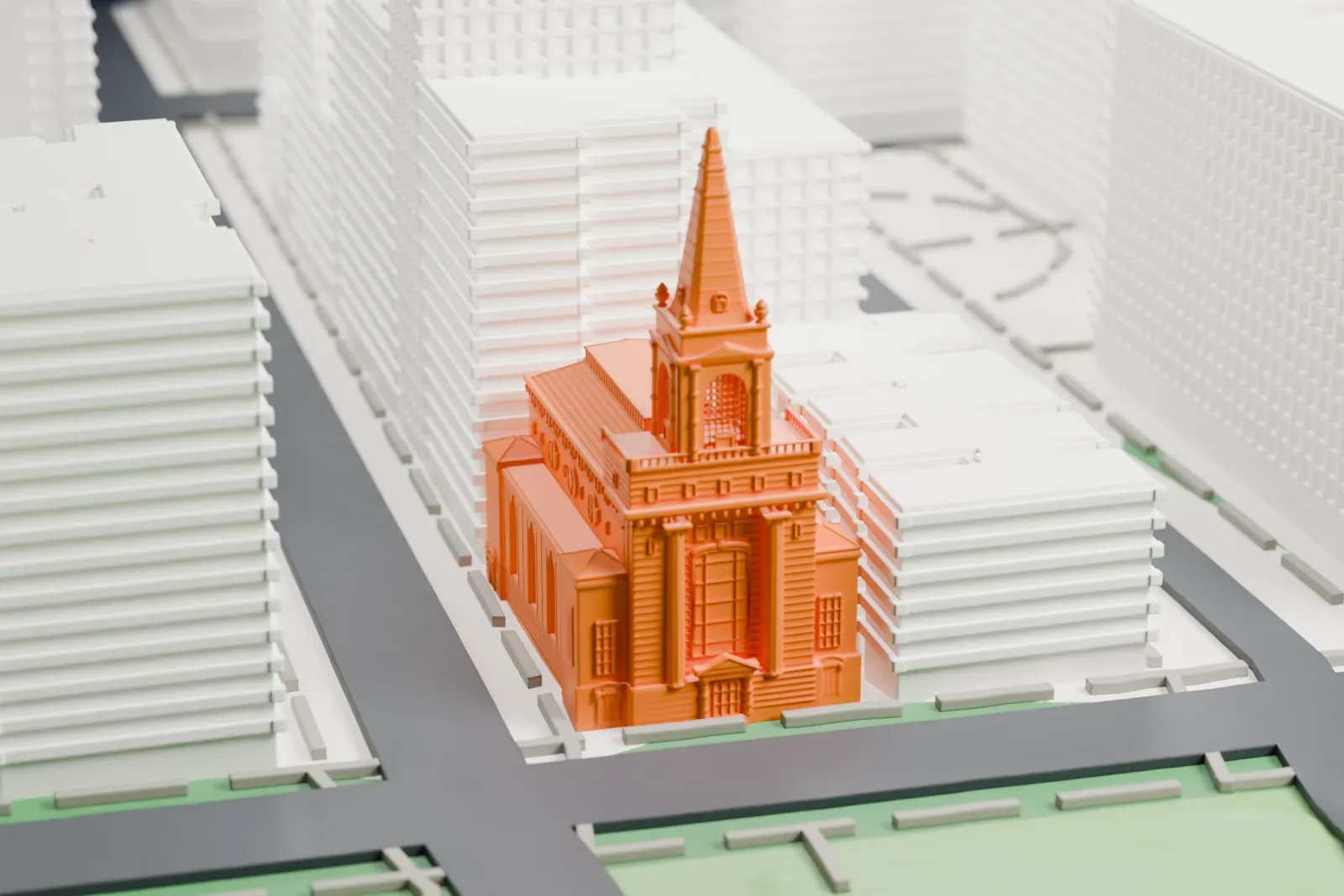 A white 3D print of First Church of Christ, Scientist, a church with a tall pointed spire on its tower, standing beside a green park with larger buildings around it.