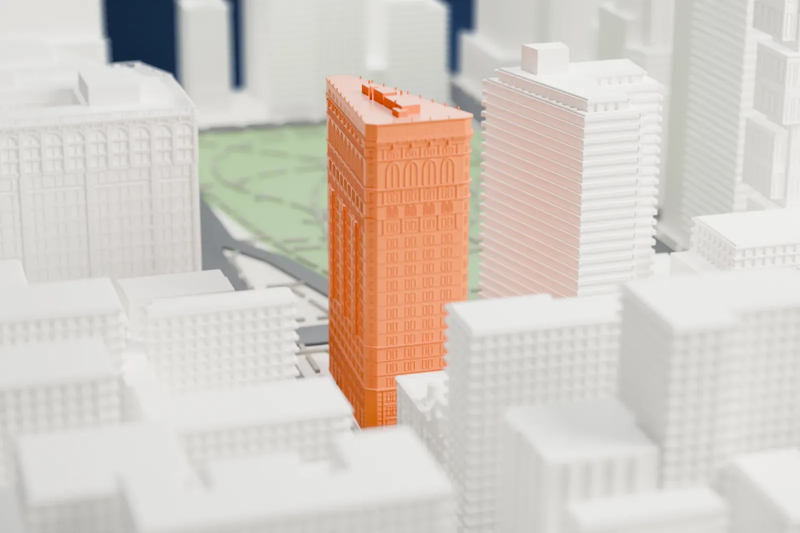 A white 3D print of the Flatiron Building, a tall triangular tower with arched windows and a narrow rounded prow, beside a green park among Manhattan blocks.