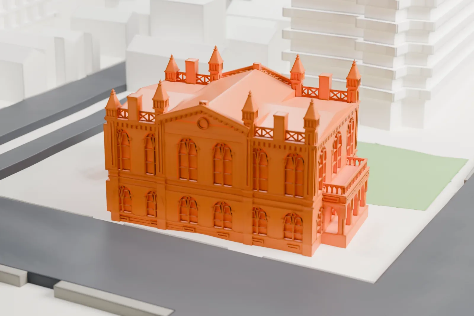 A white 3D print of Flushing Town Hall, a small brick building with corner turrets and a multi-slope roof, beside a green lawn, a street and taller buildings.