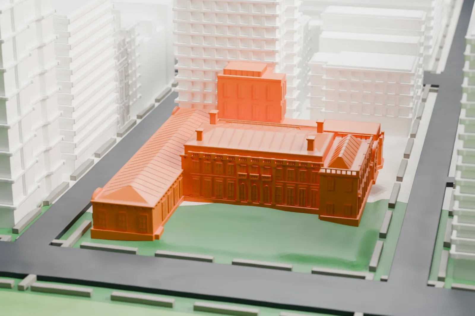 A white 3D print of the Frick Collection's mansion, a long building with hipped roofs and wings around a green lawn, with tall buildings and a park nearby.
