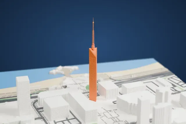 A white 3D print of Fukuoka Tower, a tall triangular tower with a thin lattice mast on top, standing among roads and low buildings near the shoreline.