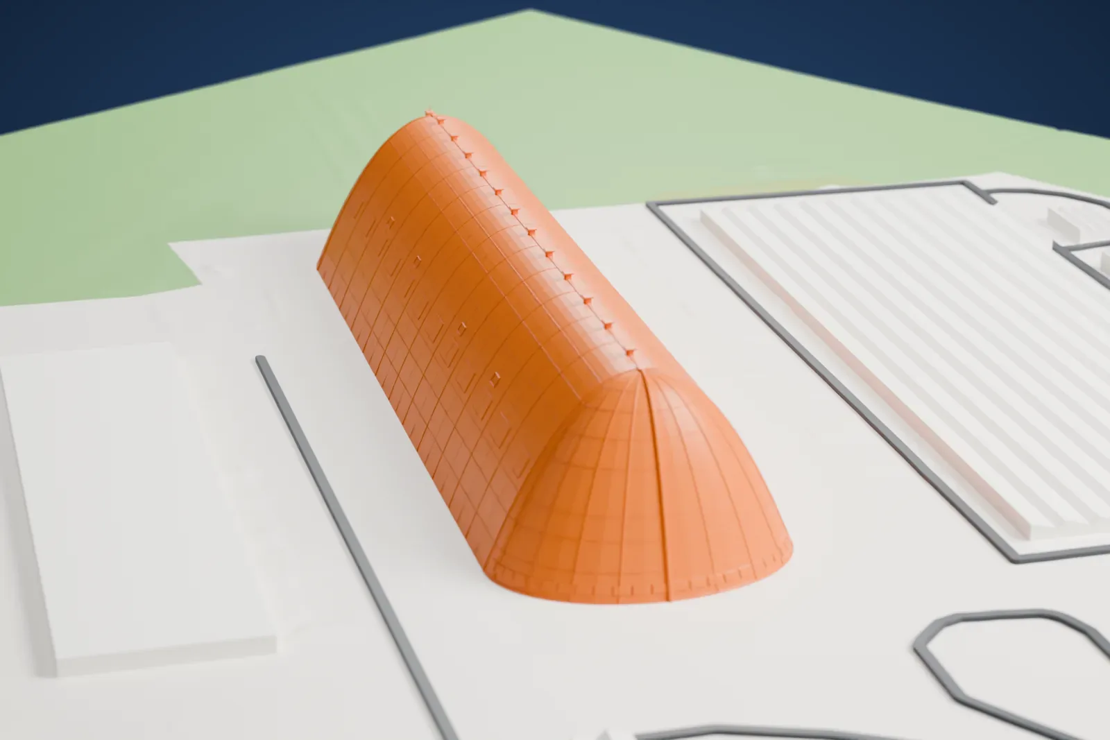 A white 3D print of the Goodyear Airdock, a long rounded hangar, standing on open ground with flat-roofed buildings and green fields around it.