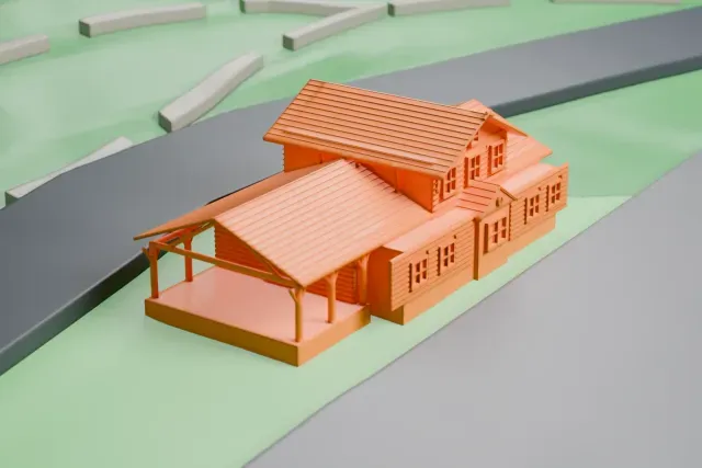 A white 3D print of the Grand Canyon Depot, a small log building with broad gabled roofs, an upper story that overhangs the first and a porch on thin posts, set on grassy ground beside wide paved areas.