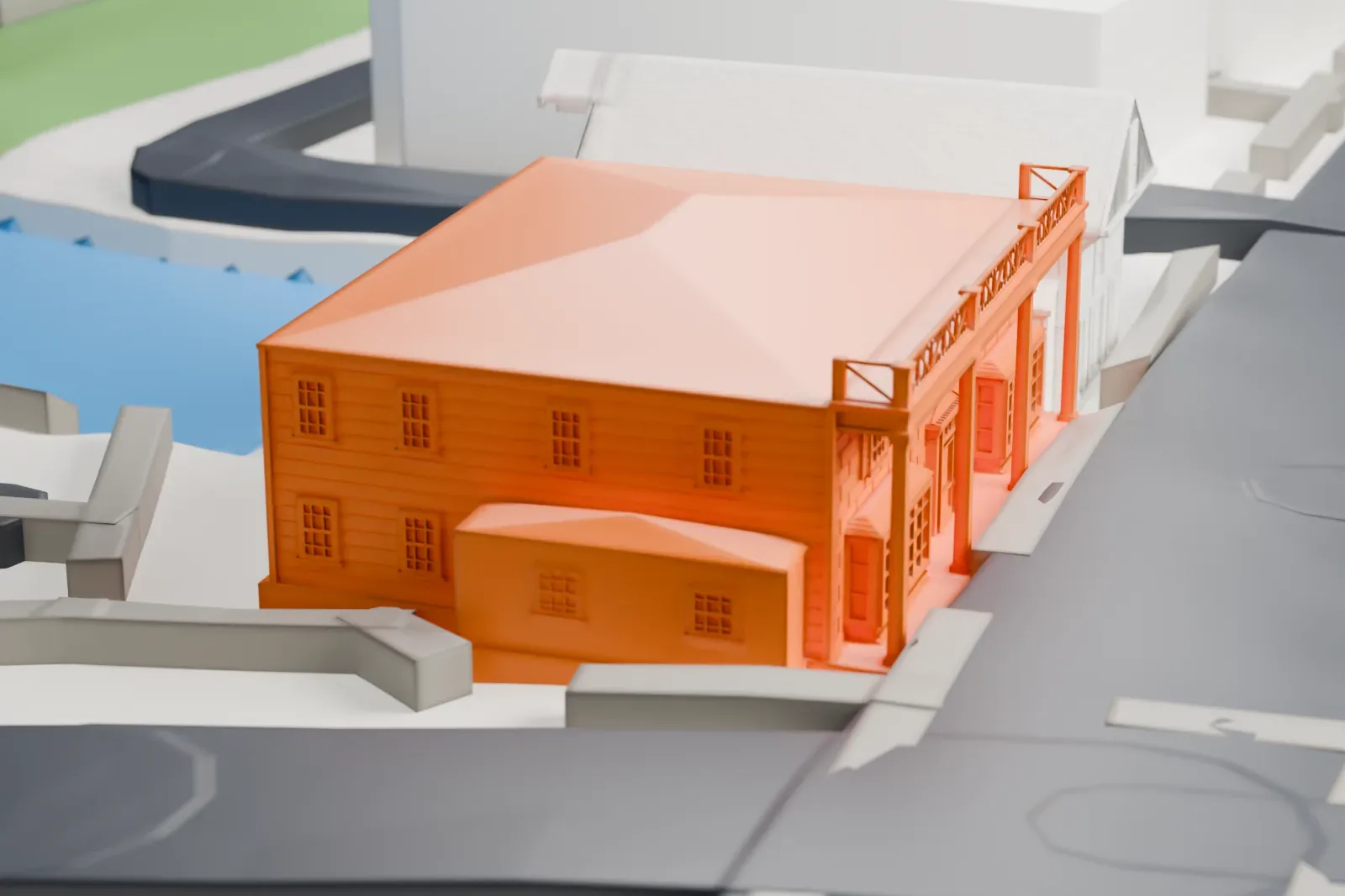 A white 3D print of the Hicks Lumber Company Store, a two-story building with a hipped roof and a front portico, beside roads, a neighboring building and a small body of water.