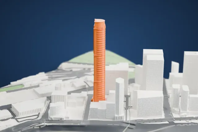 A white 3D print of Infinity Tower, a very tall, slender tower with a rounded profile and horizontal bands, rising among lower buildings near a wide stretch of water.
