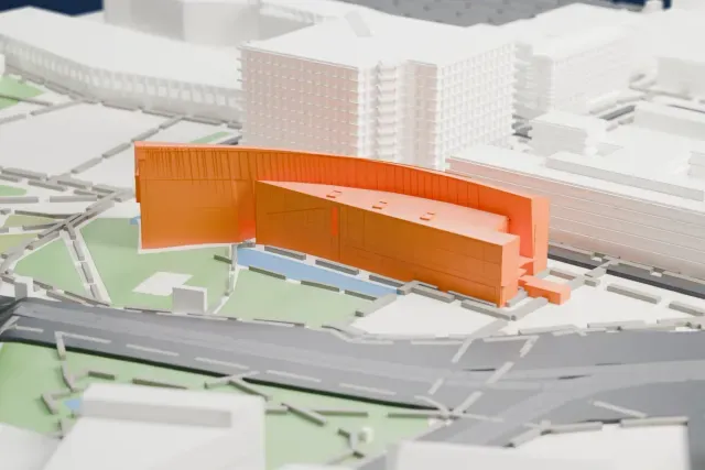 A white 3D print of Kiasma, a long curved building with a ribbed shell and a glazed front, beside a park and a pool, among roads and larger buildings.