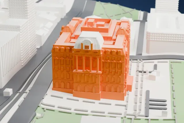 A white 3D print of the Libertad Palace, a large block-shaped palace with ornate facades, a domed central pavilion with a clock face and domed corner pavilions, beside wide roads, parkland and neighboring towers.