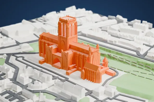 A white 3D print of Liverpool Cathedral, a long Gothic church with a tall square central tower, standing on a green slope with streets and buildings around it.