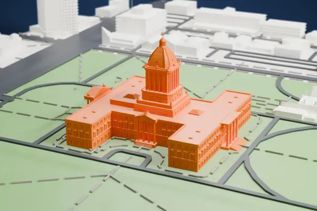 A white 3D print of the Manitoba Legislative Building, a cross-shaped building with a central dome, on green lawns with curving roads.