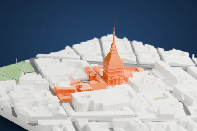 A white 3D print of the Mole Antonelliana, a tall domed tower with a very slender spire, standing among the dense gray buildings of Turin.