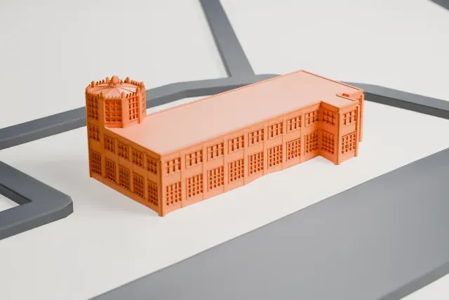 A white 3D print of the Old Port Columbus Terminal, a long, low building with rows of windows and a many-sided tower at one end topped by a toothed crown, on open ground between roads.