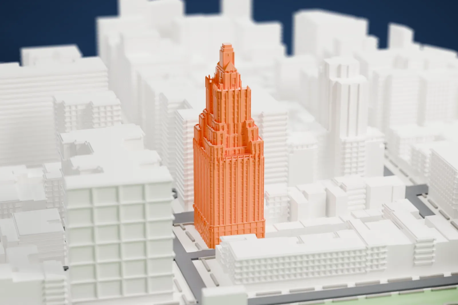 A white 3D print of One Fifth Avenue, a tall tower that narrows in stepped stages to a small pyramid-capped top, standing beside a green park among lower buildings.