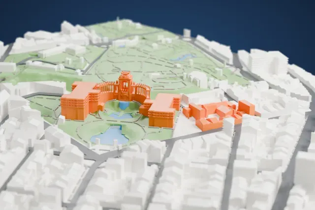 A white 3D print of the Palais Longchamp, an ornate central structure with a curved colonnade, set in green parkland with ponds and surrounded by gray buildings.