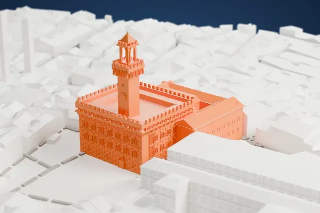 A white 3D print of the Palazzo Vecchio, a block-shaped palace with battlements and a tall slender tower, among densely packed buildings with an open square beside it.