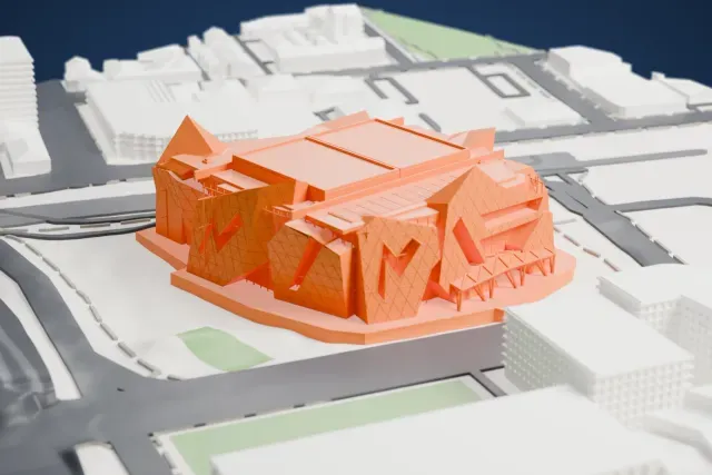 A white 3D print of Perth Arena, a faceted venue with a diamond-lattice facade and a ridged roof, standing on a low base among roads, grassy patches and nearby buildings.