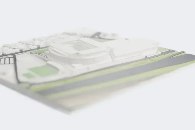 A white 3D print of RCDE Stadium, a round football stadium with a grid-like roof and corner trusses, standing on open ground beside roads and large low buildings.