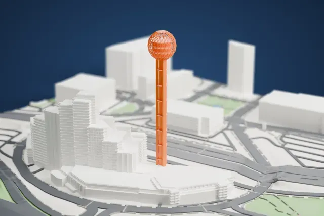 A white 3D print of Reunion Tower, a tall slender shaft topped by an open lattice sphere, standing among buildings, wide roads and patches of park.