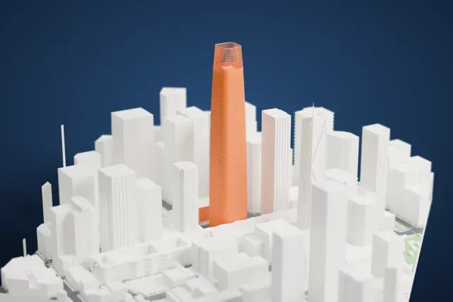 A white 3D print of Salesforce Tower, a tall tapering tower with an open lattice crown, rising above a dense cluster of downtown buildings.