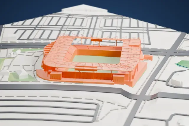 A white 3D print of Sporting Park, an open soccer stadium with a green pitch, tiered stands and a thin roof frame, surrounded by roads, parking areas, small buildings and patches of green.