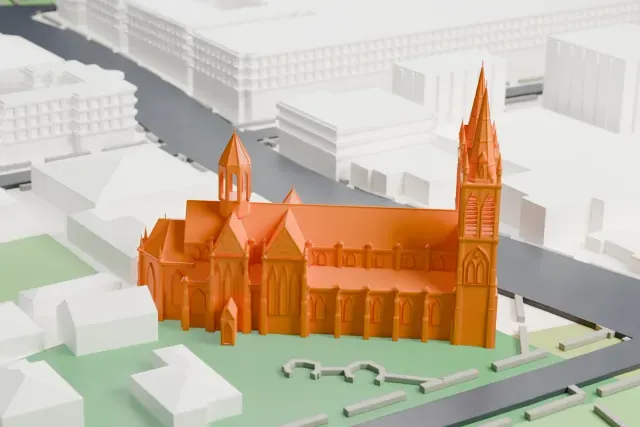 A white 3D print of St Peter's Cathedral, a Gothic church with two tall spired towers and steep roofs, standing on green lawns beside roads and low buildings.