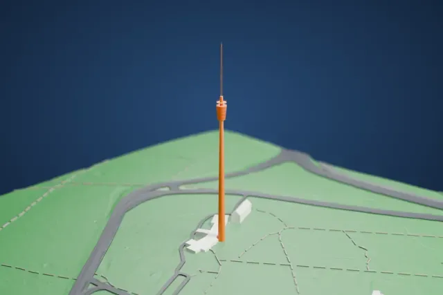 A white 3D print of Fernsehturm Stuttgart, a very slender tower with a round basket near the top and a thin antenna mast, standing on green parkland with a few low buildings at its base.