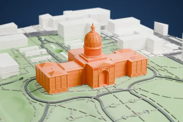 A white 3D print of the Texas State Capitol, a symmetrical building with a ribbed dome and slim spire, a columned portico and pyramid-roofed corner wings, standing on lawns with curving drives and downtown buildings around it.