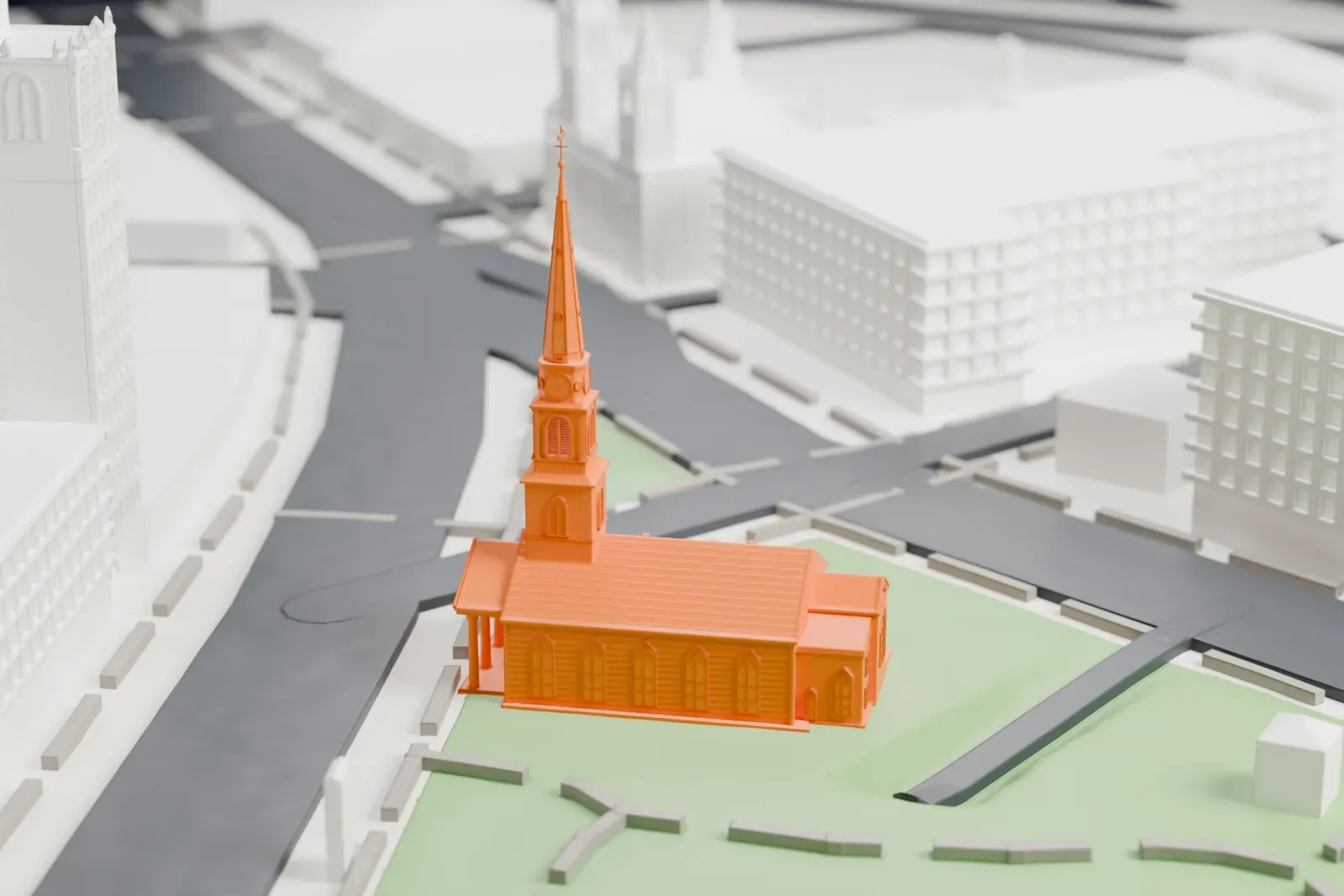 A white 3D print of Trinity & St. Philip's Cathedral, a church with a tall tapering spire, standing in a green park beside streets and larger buildings.