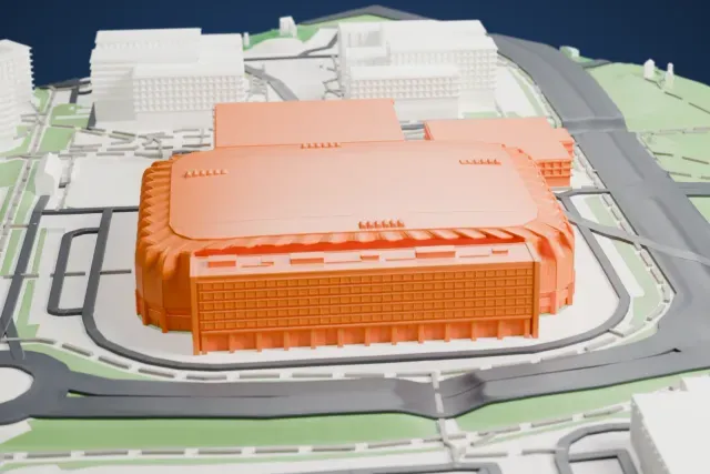A white 3D print of Unity Arena, a wide indoor arena with a flat roof, a ribbed edge and a taller wing along one side, among roads and neighboring buildings.