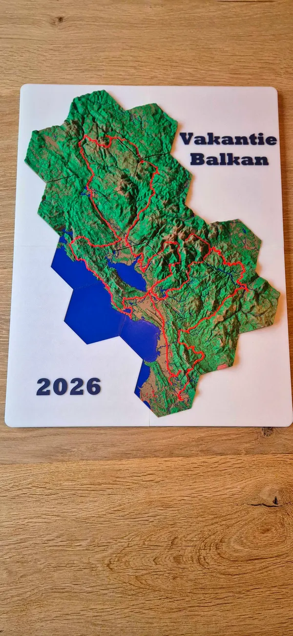 3D printed terrain map of the Balkans cut into hexagon tiles, green mountains with a red route and blue sea, on a white plate that reads Vakantie Balkan 2026.