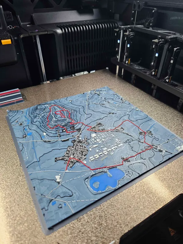 3D printed terrain map in blue-gray with contour lines, a small town, a blue lake and a route in red over the hills, still on the printer bed.