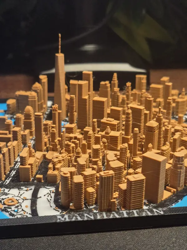 3D printed map of Lower Manhattan with tan skyscrapers on black streets and blue water, the name Manhattan on the frame.