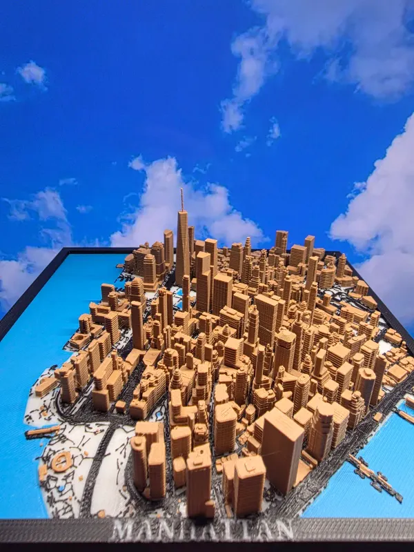 3D printed Lower Manhattan skyline in tan filament in a black frame, photographed against a blue sky backdrop.