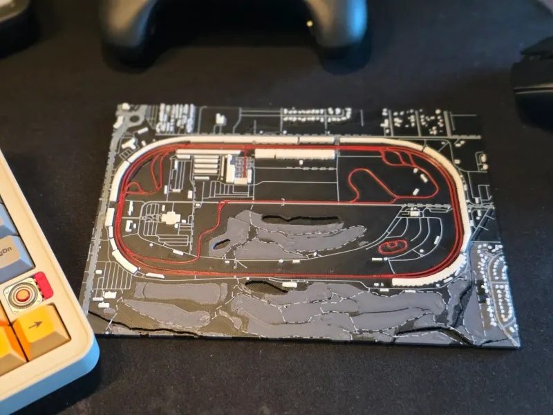 3D printed map of an oval racetrack in dark gray with the lap in red, lying on a desk.