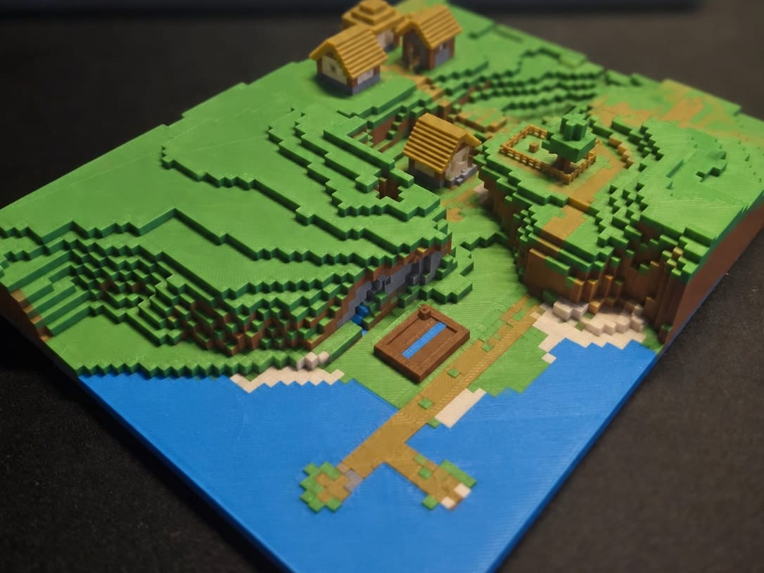 A 3D-printed multicolor Minecraft village with terraced grass hills, houses, a dock, and blue water, exported from a Java world with Mini Skyline.