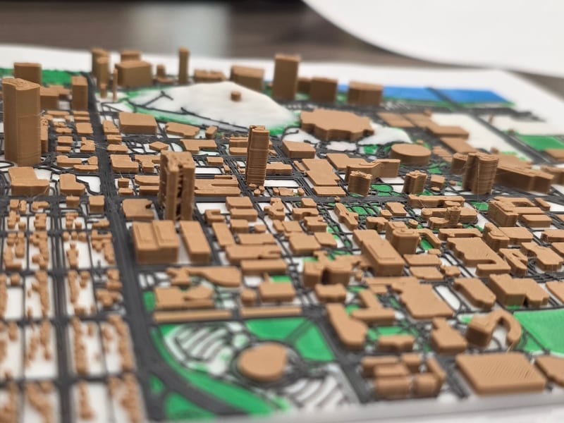 3D printed city map with tan buildings, dark roads, green parks, and blue water.